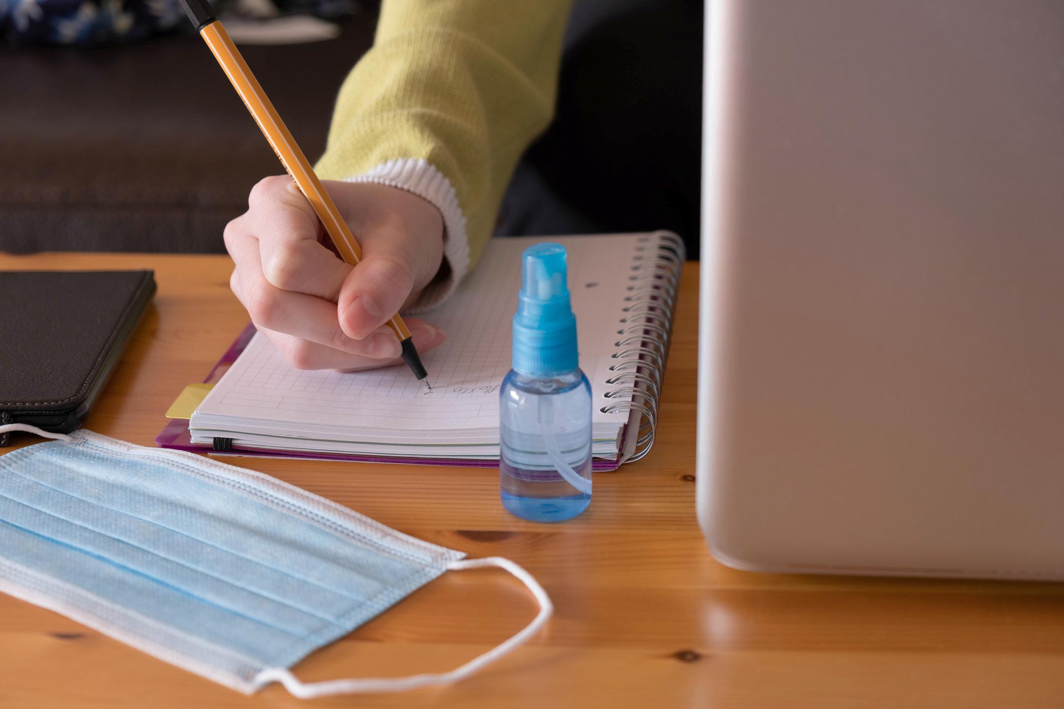 student writing notes with mask and hand sanitizer on table